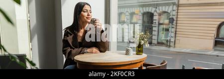 brunette femme en manteau boire du café et regarder par la fenêtre dans le café de prague, bannière, image de stock