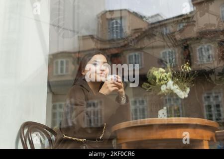 jeune femme brunette en manteau buvant du café à partir d'une coupe de papier près de la fenêtre dans le café de prague, image de stock