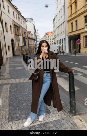 Femme élégante en manteau buvant un café d'une tasse de papier dans la rue à Prague, image de stock Banque D'Images