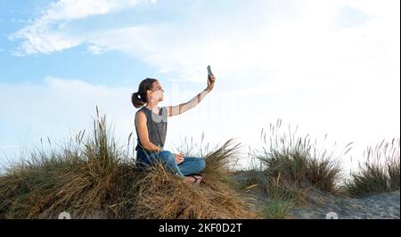 Femme caucasienne assise sur une dune de sable prenant un selfie sur un téléphone portable. Banque D'Images
