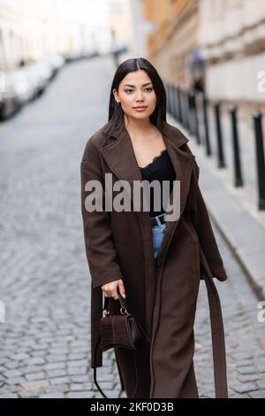 femme brune élégante en manteau brun tenant sac à main tout en marchant dans la rue à prague, image de stock Banque D'Images