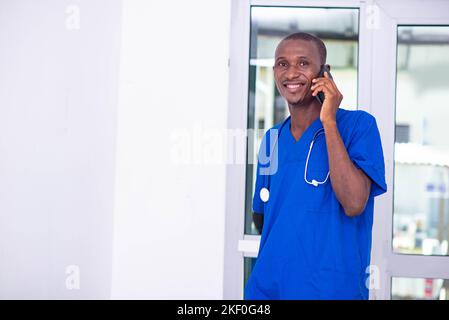 beau jeune homme médecin en uniforme bleu avec stéthoscope parlant sur téléphone portable à l'hôpital tout en souriant. Banque D'Images