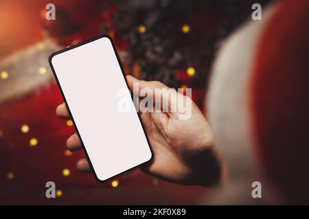 homme avec chapeau de père noël tenant téléphone mobile à la main avec écran blanc sur fond de décorations de noël. achats de cadeaux en ligne et publicité Banque D'Images