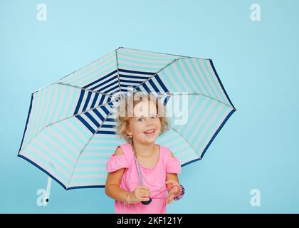 Petite fille gaie se tenir sous un parapluie ouvert sur fond bleu vide, espace libre de copie. L'enfant heureux de la maternelle prend refuge de la pluie. La vie Banque D'Images