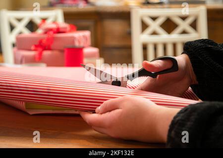 Femme emballant un cadeau en papier strié avec un ruban rouge. Mains d'emballage d'une boîte dans le papier rouge et blanc de noël. Banque D'Images