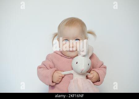 Mignon bébé fille jouer avec animal bourré, isolé sur blanc. Un tout-petit souriant se tient avec plaisir à la main, le lapin en peluche. Enfant blond aux cheveux vêque de vêtements roses Banque D'Images