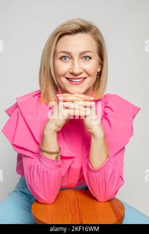 Adorable femme adulte avec maquillage parfait de levage dans un chemisier rose vif assis avec chaise, isolé sur blanc. Portrait d'un succès assuré Banque D'Images