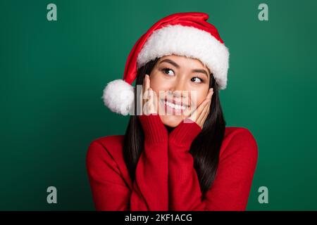 Portrait photo de jeune torride beaming sourire chinois fille porter santa Hat toucher joues rêve regard vide espace isolé sur fond vert couleur Banque D'Images