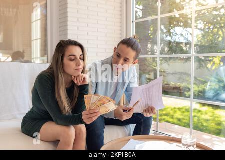 Les jeunes couples se préoccupent de l'argent restant pour le mois, regardant des factures de 50 euros entre les mains. Assis dans le canapé Banque D'Images