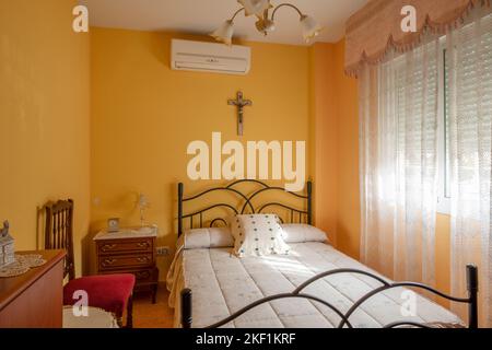 Chambre avec un petit lit avec un cadre en métal noir, des tables de chevet en bois, des murs peints en jaune et un crucifix sur le mur Banque D'Images