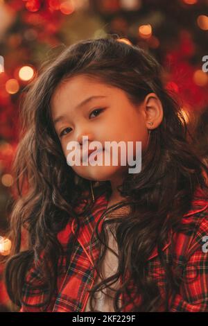 Bonne petite fille de rêve de japon regardant loin en attendant miracle de Santa posant près de l'arbre de noël. Asiatique Kid noir cheveux bouclés dans un costume rouge à carreaux fête Banque D'Images