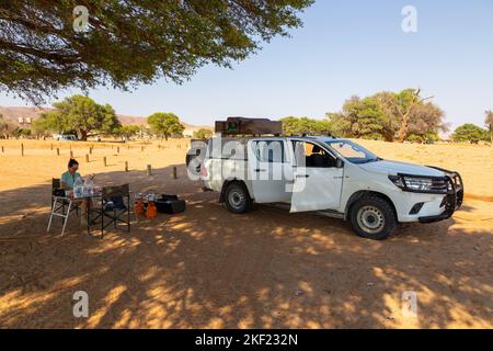 Camping Sesriem, Sossusvlei, Namibie - 01 octobre 2018 : une fille assise sous un acacia au camping. Voiture avec équipement garé à proximité. Banque D'Images
