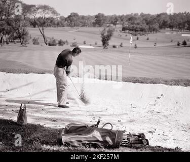 1950S HOMME FRAPPANT LA BALLE DE GOLF HORS D'UN PIÈGE À SABLE DANGEREUX SUR LE SAC VERT ET LES CLUBS SUR LA POSITION ALLONGÉE TERRAIN DERRIÈRE LUI - G451 HAR001 HARS STRATÉGIE DE GOLFEURS ET OBSTACLE DE LOISIRS DE LA DIRECTION DU PIÈGE DE LA VUE ARRIÈRE VERS LE HAUT SUR LE PIÈGE DE SABLE MOTION FLOU CONCEPTUEL DE DERRIÈRE LES LIENS D'ÉCHAPPEMENT VUE ARRIÈRE LES DANGERS LUI ADULTE MOYEN-ADULTE FEMMES SOLUTIONS NOIR ET BLANC BUNKER CAUCASIEN ETHNICITÉ HAR001 ANCIENNE Banque D'Images