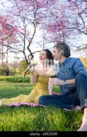 Un couple sur une couverture de pique-nique à carreaux dans Forest Park avec une bouteille de vin et des bud-arbres rouges derrière eux. Banque D'Images