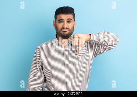 Portrait d'un jeune homme d'affaires adulte critiquant la mauvaise qualité avec des pouces vers le bas Grimace déplaisée, montrant le geste déplaît, portant une chemise rayée. Studio d'intérieur isolé sur fond bleu. Banque D'Images