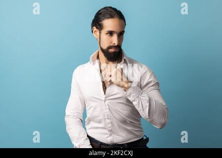 Portrait d'un homme beau et sérieux avec une barbe vêtue d'une chemise blanche, regardant loin de l'expression du visage concentrée, en gardant la main sur le collier. Studio d'intérieur isolé sur fond bleu. Banque D'Images