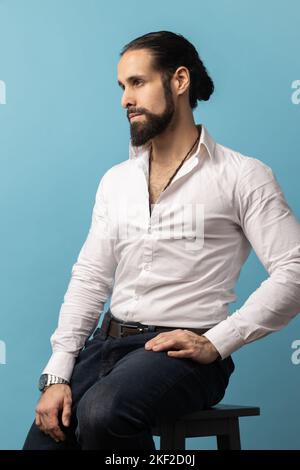 Vue latérale d'un beau homme avec barbe et cheveux foncés recueillis en portant une chemise blanche et un pantalon noir, assis et regardant loin avec l'expression pensive. Studio d'intérieur isolé sur fond bleu Banque D'Images