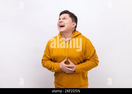 Portrait d'un homme heureux extrêmement excité tenant son ventre et en riant à voix haute, en riant à rire de plaisanterie amusante, portant un sweat à capuche de style urbain. Studio d'intérieur isolé sur fond blanc. Banque D'Images