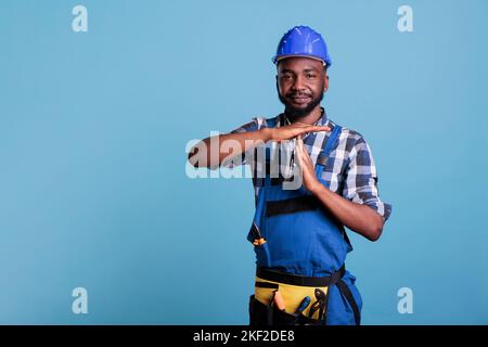 Travailleur de la construction faisant le geste de pause sur la caméra, montrant le signe d'arrêt en forme de T avec les bras dans le studio. Un constructeur afro-américain qui demande un temps d'arrêt pour une courte pause, portant un casque et une combinaison. Banque D'Images