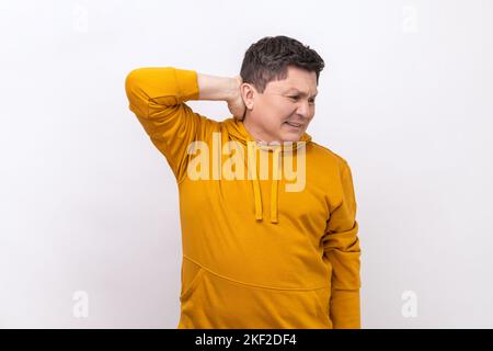 Inquiet homme anxieux touchant le cou, sensation de douleur aiguë se déplaçant et tournant la tête, souffrant des problèmes de colonne vertébrale, ostéochondrose, port de sweat à capuche de style urbain. Studio d'intérieur isolé sur fond blanc. Banque D'Images