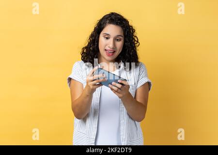 Loisirs et technologie. Femme utilisant un téléphone portable avec une expression stupéfaite, jouant à des jeux vidéo sur le téléphone portable, bavardant sur les réseaux sociaux. Studio d'intérieur isolé sur fond jaune. Banque D'Images