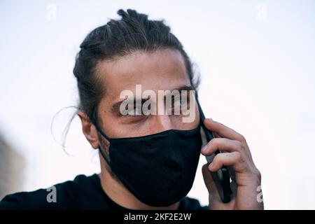 caucasien mec noir t-shirt disheveled cheveux longs avec masque de visage sur la rue parlant sur son smartphone dans la ville Banque D'Images