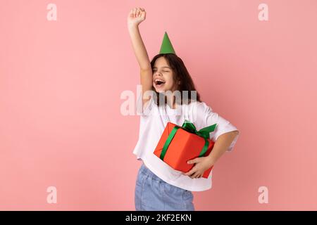 Portrait d'une petite fille portant un T-shirt blanc et avec un cône de fête tenant une boîte cadeau et criant pour la joie, célébrant l'anniversaire, réjouissant le présent. Studio d'intérieur isolé sur fond rose. Banque D'Images