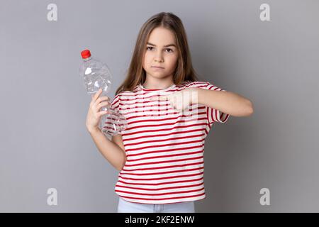 Portrait de sérieuse charmante petite fille portant un T-shirt rayé tenant et pointant le doigt sur la bouteille en plastique vide, regardant l'appareil photo. Prise de vue en studio isolée sur fond gris. Banque D'Images