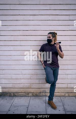 homme caucasien avec de longues cheveux de barbe debout dans le t-shirt noir penché sur le mur sale de ville avec smartphone dans la main gauche portant un masque facial Banque D'Images