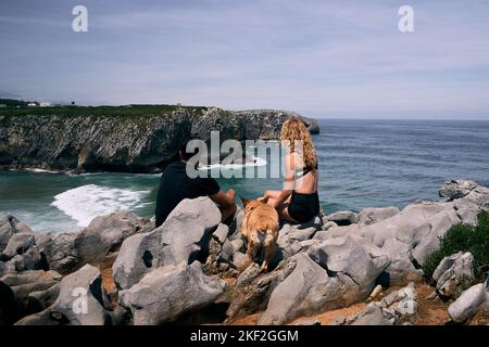 latino garçon et blonde caucasienne fille assise sur les grands rochers à côté de leur chien contemplant les vagues de la mer, ribadesella asturias, espagne Banque D'Images