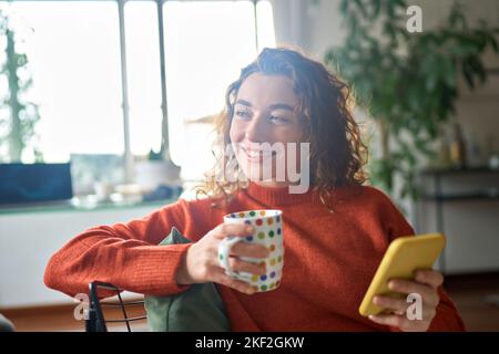 Jeune femme heureuse utilisant un téléphone mobile boire un café rêvant à la maison. Banque D'Images
