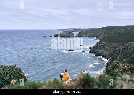 latino garçon assis sur le rocher avec une veste jaune contemplant la beauté de la côte et des falaises, cabo de penas, espagne Banque D'Images