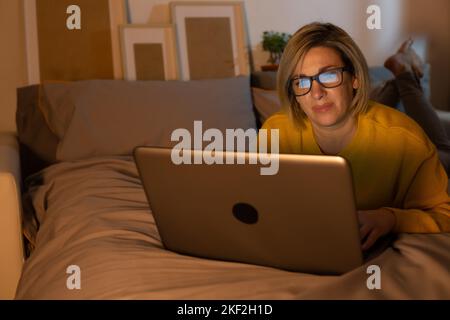 Belle femme caucasienne de 40 ans en lunettes utilise et travaille sur un ordinateur portable avec un écran lumineux tard dans la nuit à la maison sur le lit. Bureau à domicile Banque D'Images