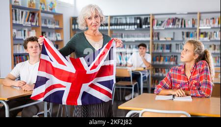 Un professeur âgé montre le drapeau de la Grande-Bretagne aux adolescents et parle de ce pays pendant la leçon à l'école de bibliothèque Banque D'Images