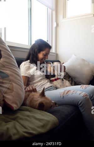 Femme hispanique assise sur un canapé riant et jouant avec ses chiens. Belle maison Banque D'Images