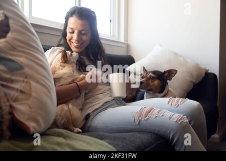 Femme hispanique assise sur un canapé riant et jouant avec ses chiens. Belle maison Banque D'Images