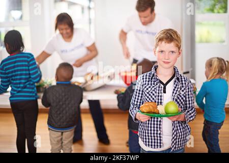 Faire une différence dans la vie des enfants. Portrait d'un petit garçon avec une assiette de nourriture avec des bénévoles servant de la nourriture en arrière-plan. Banque D'Images