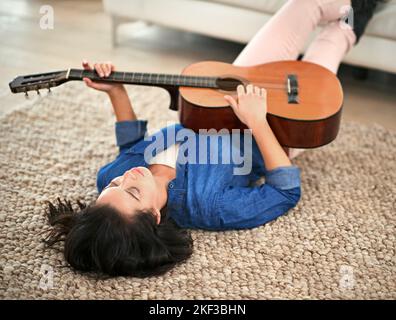 La musique est ce à quoi ressemblent les sentiments. Une jeune femme jouant de la guitare en étant allongée sur le sol de son salon. Banque D'Images