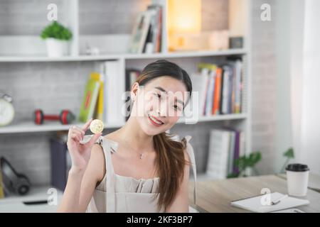 Femme asiatique souriante tenant la chaîne de blocs de crypto-monnaie en bitcoin or sur table en bois bureau dans la salle de séjour à la maison. Crypto monnaie blockchain d'économie Banque D'Images