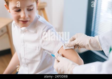 Gros plan d'une prise de vue d'une femme médecin méconnue qui vaccine un petit garçon sur l'épaule à l'hôpital par fenêtre. Banque D'Images