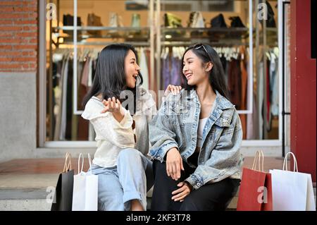 Deux jeunes femmes asiatiques surjoyeuses et heureuses aiment parler et rire tout en s'asseyant sur les escaliers en face du magasin de clothe ensemble Banque D'Images