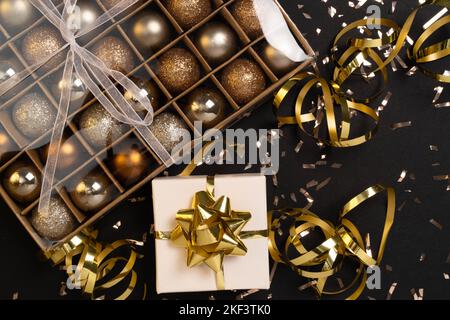 Boîte cadeau de Noël ou cadeau avec ruban, confettis dorés et boules dorées sur fond noir. Carte de vœux du nouvel an magique. Décoration de Noël. Maquette. Pose à plat. Banque D'Images