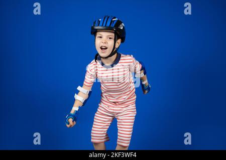 Un joyeux garçon dans un casque de protection, des protège-coudes et des gants sur fond bleu. Protection lors de la conduite d'un vélo, d'un skateboard ou d'un patin à roulettes Banque D'Images