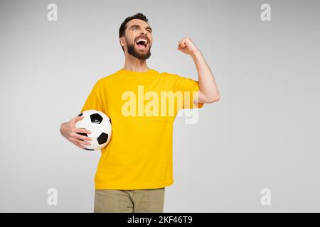 fan de sport débord de joie avec ballon de football regardant loin et montrant le geste de gain tout en criant isolé sur gris, image de stock Banque D'Images
