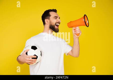 fan de sport barbu en t-shirt blanc tenant le ballon de football et criant en mégaphone isolé sur jaune, image de stock Banque D'Images