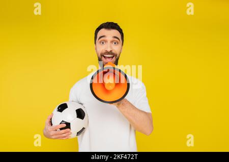 fan de sport stupéfié avec le ballon de football criant au mégaphone et regardant la caméra isolée sur jaune, image de stock Banque D'Images