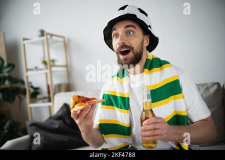 un homme émerveillé dans un chapeau tenant une tranche de pizza savoureuse et une bouteille de bière tout en regardant le championnat, image de stock Banque D'Images