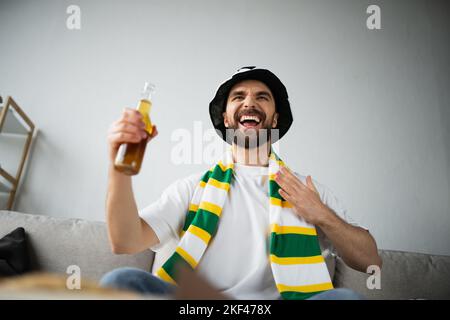 vue en contre-plongée d'un fan de sport stupéfié dans un foulard et un chapeau tenant une bouteille de bière tout en regardant le championnat, image de la bourse Banque D'Images