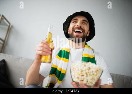 fan de sport positif dans un chapeau et un foulard tenant un bol avec pop-corn et une bouteille de bière tout en regardant le championnat, image de stock Banque D'Images