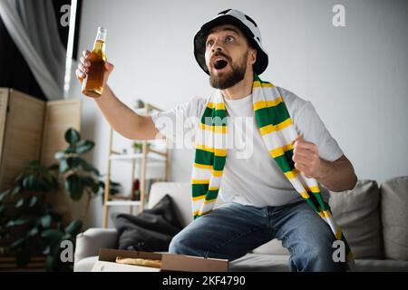 homme à barbe excité dans un chapeau de fan sportif et une écharpe tenant une bouteille de bière tout en regardant le championnat, image de stock Banque D'Images